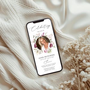 May include: A smartphone displays a memorial invitation with the text "Celebrating the Life of Lynn Hutchins." The design features a photo of a woman surrounded by floral accents in shades of pink and purple. The phone rests on a cream-colored surface.