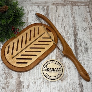 May include: A wooden bread slicer with a light wood cutting board and a darker wood frame. A wooden handle with a stainless steel blade is positioned above the board. A wooden coaster with the text "Spencer Wood Creations" is also present.