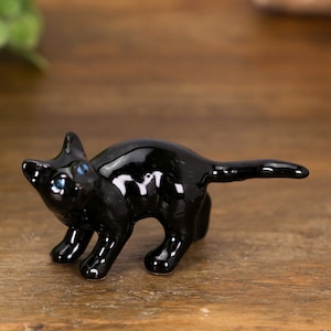 May include: A small, glossy black ceramic cat figurine with bright blue eyes, depicted in a standing position with its back arched. The figurine is positioned next to a gold-colored coin for scale, highlighting its miniature size and smooth finish.