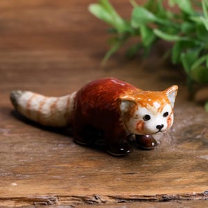 May include: A small, glossy ceramic figurine of a red panda. The animal features a deep reddish-brown body, a white face with dark eye markings, and a long, bushy tail with alternating light and dark brown rings. It stands on four dark paws.