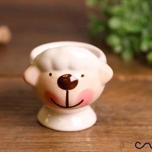 Puede incluir: Un huevero de cerámica con forma de oveja, blanco con mejillas rosas, nariz y boca marrones. El huevero está sobre una superficie de madera.