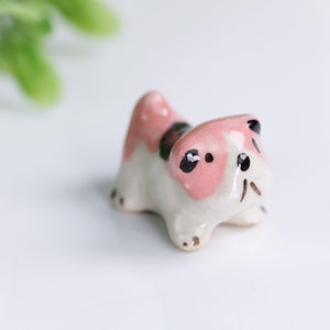Könnte beinhalten: Eine kleine Keramikfigur eines Hundes mit rosa und weißer Farbe. Der Hund hat schwarze Augen und eine schwarze Nase.