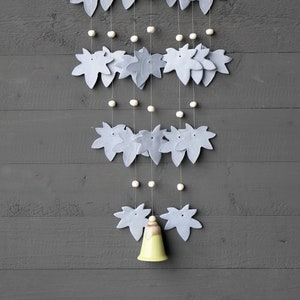 May include: A decorative wall hanging with light blue leaf shapes, small white beads, and a yellow bell. The design is suspended from a natural wood branch. The background is a grey wooden wall. The overall aesthetic is minimalist.