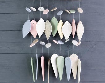 May include: A hanging mobile featuring a horizontal wooden dowel suspended by white string. Attached are multiple thin, dangling ceramic pieces in various pastel shades including blue, pink, green, yellow, and white, shaped like leaves and small circles.