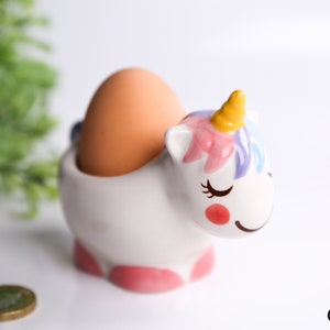 Può includere: Un porta uova in ceramica bianca a forma di unicorno con dettagli rosa, viola e gialli. L'unicorno ha gli occhi chiusi e una bocca sorridente. Un uovo è seduto nel porta uova.