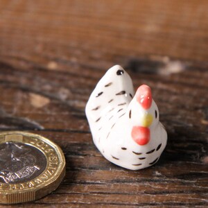 Puede incluir: Una peque&ntilde;a figurita de pollo de cer&aacute;mica blanca con marcas negras y acentos rojos y amarillos. La figurita est&aacute; sentada sobre una superficie de madera.