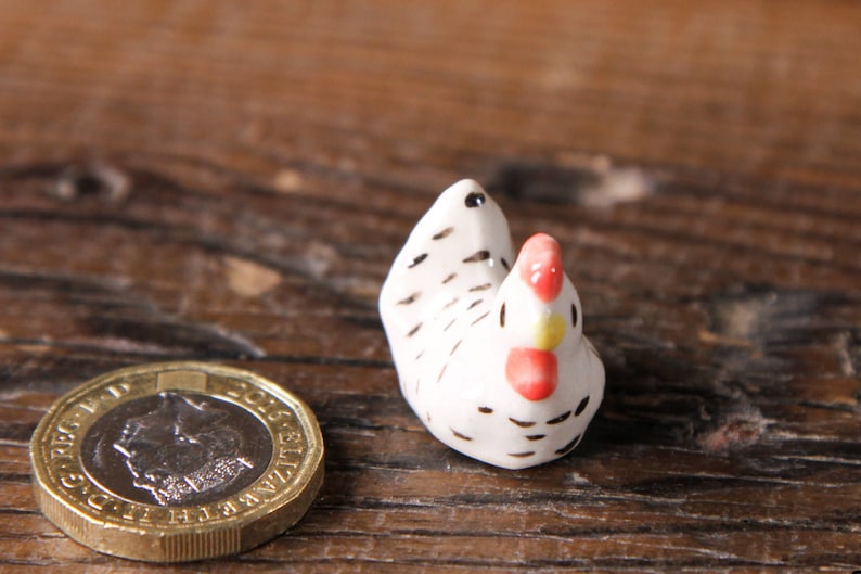 Puede incluir: Una peque&ntilde;a figurita de pollo de cer&aacute;mica blanca con marcas negras y acentos rojos y amarillos. La figurita est&aacute; sentada sobre una superficie de madera.