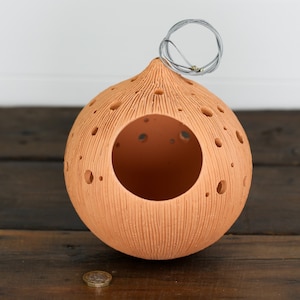 Puede incluir: Una casita de pájaros de terracota con forma de esfera con una gran abertura y un lazo de alambre en la parte superior. La casita de pájaros tiene una superficie texturizada y pequeños agujeros por todas partes.