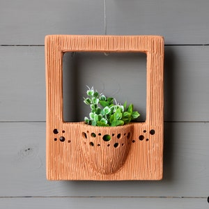 Könnte beinhalten: Ein Wandbepflanzer aus Terrakotta mit einem quadratischen Rahmen und einem runden, durchbrochenen Topf im Inneren. Der Pflanzer hat eine strukturierte Oberfläche und ist mit kleinen Löchern verziert. Eine kleine grüne Pflanze wächst im Topf.