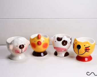 May include: A set of four ceramic egg cups featuring animal designs. The cups are shaped like a white sheep, a yellow chicken, a black and white cow, and an orange cat. Each cup has a rounded base and a wide opening for holding a single egg.