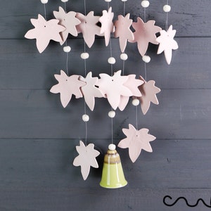 May include: A decorative mobile with pale pink leaf shapes and white beads suspended on strings. A green and brown bell hangs at the bottom. The backdrop is a dark grey wooden surface.