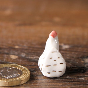 Puede incluir: Una peque&ntilde;a figurita de pollo de cer&aacute;mica blanca con rayas marrones y una cresta roja. La figurita est&aacute; sentada sobre una superficie de madera.