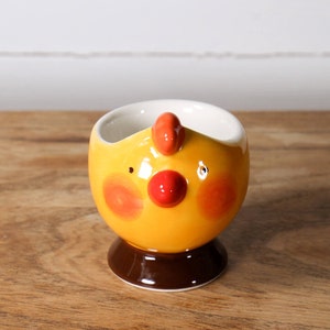 May include: A yellow ceramic egg cup shaped like a chicken with red and orange accents. The cup has a brown base.