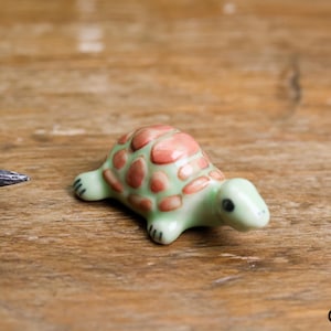May include: A small ceramic turtle figurine with a green and pink shell. The turtle is sitting on a wooden surface.
