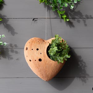 May include: A heart-shaped, terracotta-colored wall planter with a textured surface and several small holes. The planter is filled with green foliage and hangs from a thin string. Greenery is also visible in the background.