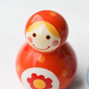 May include: A vibrant orange wooden toy with a smiling face and a flower design. The toy has a rounded shape with a smaller head and a larger body. It is decorated with polka dots and a flower.