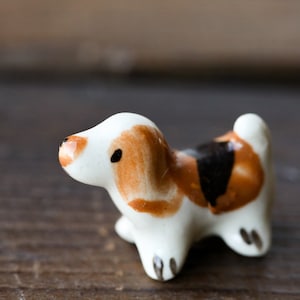 Puede incluir: Una pequeña figurita de perro de cerámica pintada en marrón, negro y blanco. El perro está de pie sobre una superficie de madera.