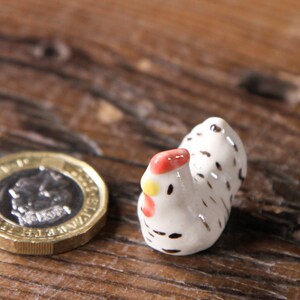 Puede incluir: Una peque&ntilde;a figurita de pollo de cer&aacute;mica blanca con manchas negras y una cresta roja. El pollo est&aacute; de pie sobre una superficie de madera.