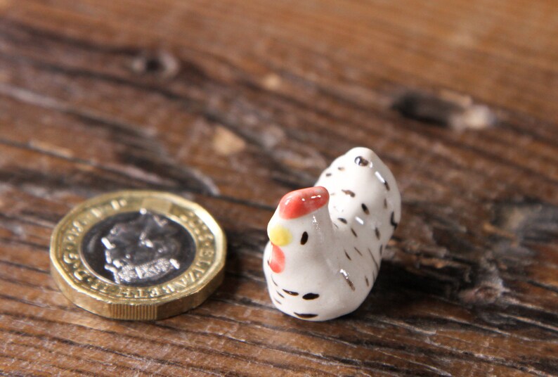 Puede incluir: Una peque&ntilde;a figurita de pollo de cer&aacute;mica blanca con manchas negras y una cresta roja. El pollo est&aacute; de pie sobre una superficie de madera.