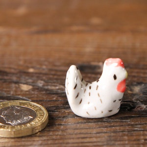 Puede incluir: Una peque&ntilde;a figurita de pollo de cer&aacute;mica blanca con manchas negras y una cresta roja. El pollo est&aacute; sentado sobre una superficie de madera.