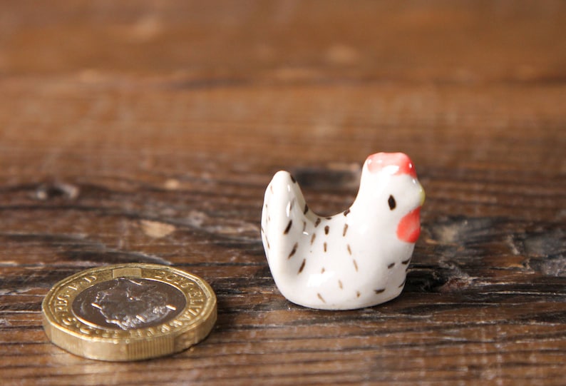 Puede incluir: Una peque&ntilde;a figurita de pollo de cer&aacute;mica blanca con manchas negras y una cresta roja. El pollo est&aacute; sentado sobre una superficie de madera.