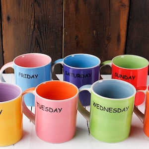 Può includere: Set di sette tazze in ceramica colorate con ogni giorno della settimana scritto su di esse. Le tazze sono disposte a semicerchio su una superficie bianca.