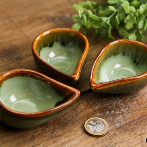 May include: Three small, teardrop-shaped ceramic bowls with a green glaze and brown rim. The bowls are arranged on a wooden surface.