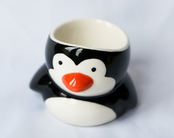 May include: A black and white ceramic egg cup shaped like a penguin with an orange beak.