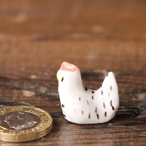 Puede incluir: Una peque&ntilde;a figurita de pollo de cer&aacute;mica blanca con manchas negras y una cresta rosa y amarilla. La figurita est&aacute; sentada sobre una superficie de madera.