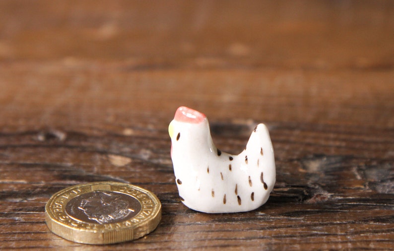 Puede incluir: Una peque&ntilde;a figurita de pollo de cer&aacute;mica blanca con manchas negras y una cresta rosa y amarilla. La figurita est&aacute; sentada sobre una superficie de madera.