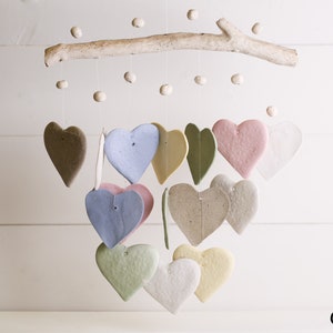Puede incluir: Móvil colgante con adornos en forma de corazón en varios colores pastel, suspendidos de una rama de madera natural. Los corazones son de color marrón, azul, rosa, verde y amarillo. Pequeñas cuentas redondas también están suspendidas.
