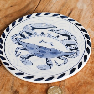May include: A blue and white ceramic trivet with a blue crab design. The trivet has a dark blue border with a rope design.