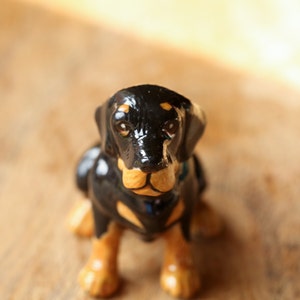 May include: A black and tan ceramic figurine of a dachshund dog sitting with its paws tucked underneath it. The dog has a black nose and brown eyes.