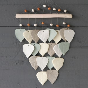 May include: A ceramic wind chime with a wooden dowel and beige, white, and light blue leaf-shaped ornaments. The wind chime is hanging from a gray wooden wall.