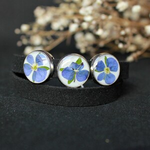 May include: A black leather bracelet with three silver round charms featuring pressed blue flowers.