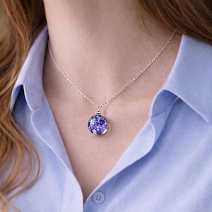 Puede incluir: Un collar de plata con un colgante redondo lleno de flores moradas. El colgante cuelga de una cadena fina. El collar se lleva sobre una camisa azul claro.