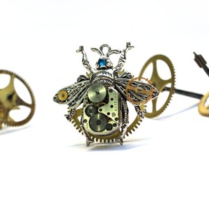 May include: A silver and gold steampunk-style insect brooch with intricate gears and a blue gemstone eye. The brooch is surrounded by smaller gears.