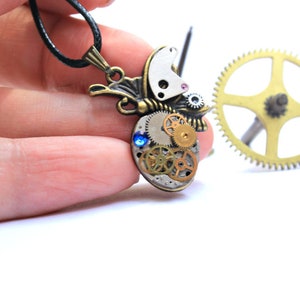 May include: A steampunk-style necklace featuring a butterfly pendant made of gears and clock parts. The pendant has a blue gemstone and is attached to a black cord.