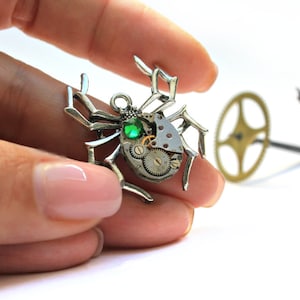 Könnte beinhalten: Ein silberner Steampunk-Spinnenanhänger mit einem grünen Edelstein in der Mitte. Die Spinne besteht aus Zahnrädern und anderen Uhrwerkteilen.