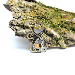 May include: A steampunk-style cat charm made of metal gears and bronze. The cat has yellow eyes and a bow tie. The charm is sitting on a piece of bark covered in moss.