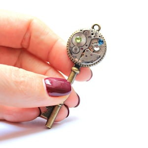 May include: A gold-toned key pendant with a round, intricate clockwork design featuring blue and green gemstones. The pendant is held in a hand with burgundy nail polish.