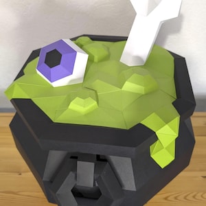 May include: A papercraft cauldron with a green, textured surface, a purple and black eye, and a white bone. The cauldron is black and sits on a wooden surface. The design is geometric and angular.