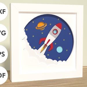 Puede incluir: Un marco blanco con un diseño de recorte de papel azul, rojo y amarillo de un cohete espacial que vuela por el espacio con estrellas y planetas.
