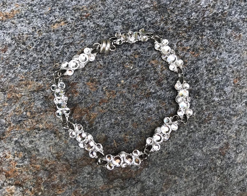 Puede incluir: Un brazalete plateado con una cadena hecha de peque&ntilde;os diamantes de imitaci&oacute;n transparentes. El brazalete tiene un cierre magn&eacute;tico.