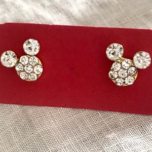 May include: A pair of gold-tone earrings with clear crystal Mickey Mouse heads. The earrings are displayed on a red background.