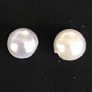 Puede incluir: Dos cuentas de perla blancas, una con un tono ligeramente blanquecino, sobre un fondo negro.