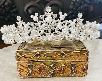 May include: A silver tiara adorned with faux pearls and clear crystals. The crown rests on a small, ornate, gold-toned box with a decorative pattern. A dark wooden clock is in the background.