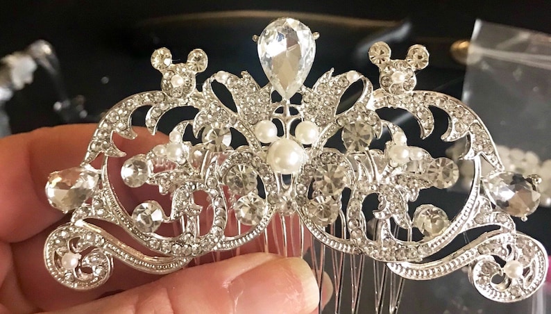 K&ouml;nnte beinhalten: Silberner Haarkamm mit einem dekorativen Design mit einem gro&szlig;en tropfenf&ouml;rmigen Kristall, kleineren Kristallen und Perlenakzenten. Das Design beinhaltet zwei Mickey Mouse Ohren auf jeder Seite.