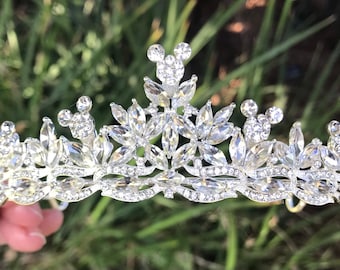 May include: A silver tiara adorned with clear crystals and Mickey Mouse motifs. The design incorporates marquise-cut crystals and round accents. A decorative headpiece for special events, it sparkles in the light.