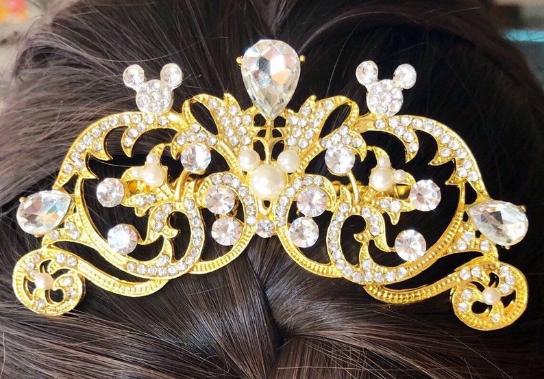 K&ouml;nnte beinhalten: Goldener Haarkamm mit einem dekorativen Design mit klaren und wei&szlig;en Strasssteinen, Perlen und Mickey-Maus-Silhouetten.